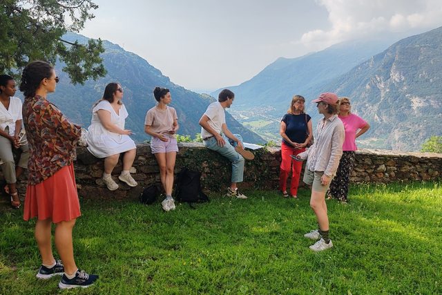 A group in conversation on a grassy terrace by a dry-stone wall, a deep alpine valley opening behind them