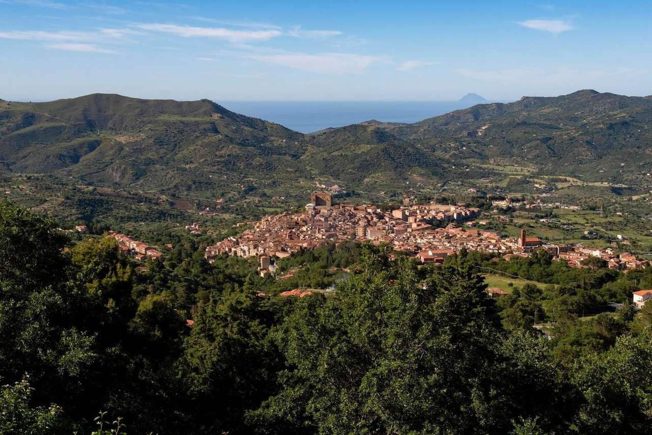 Hill town of terracotta roofs in a green valley, with wooded mountains and the sea beyond