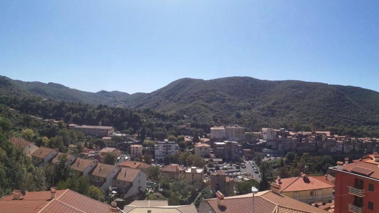 A hill town of terracotta roofs below wooded mountains under a clear blue sky