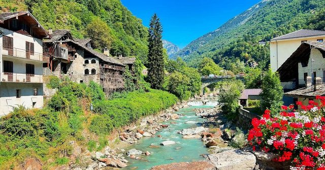 A clear alpine river between stone houses and wooded slopes, red geraniums in the foreground