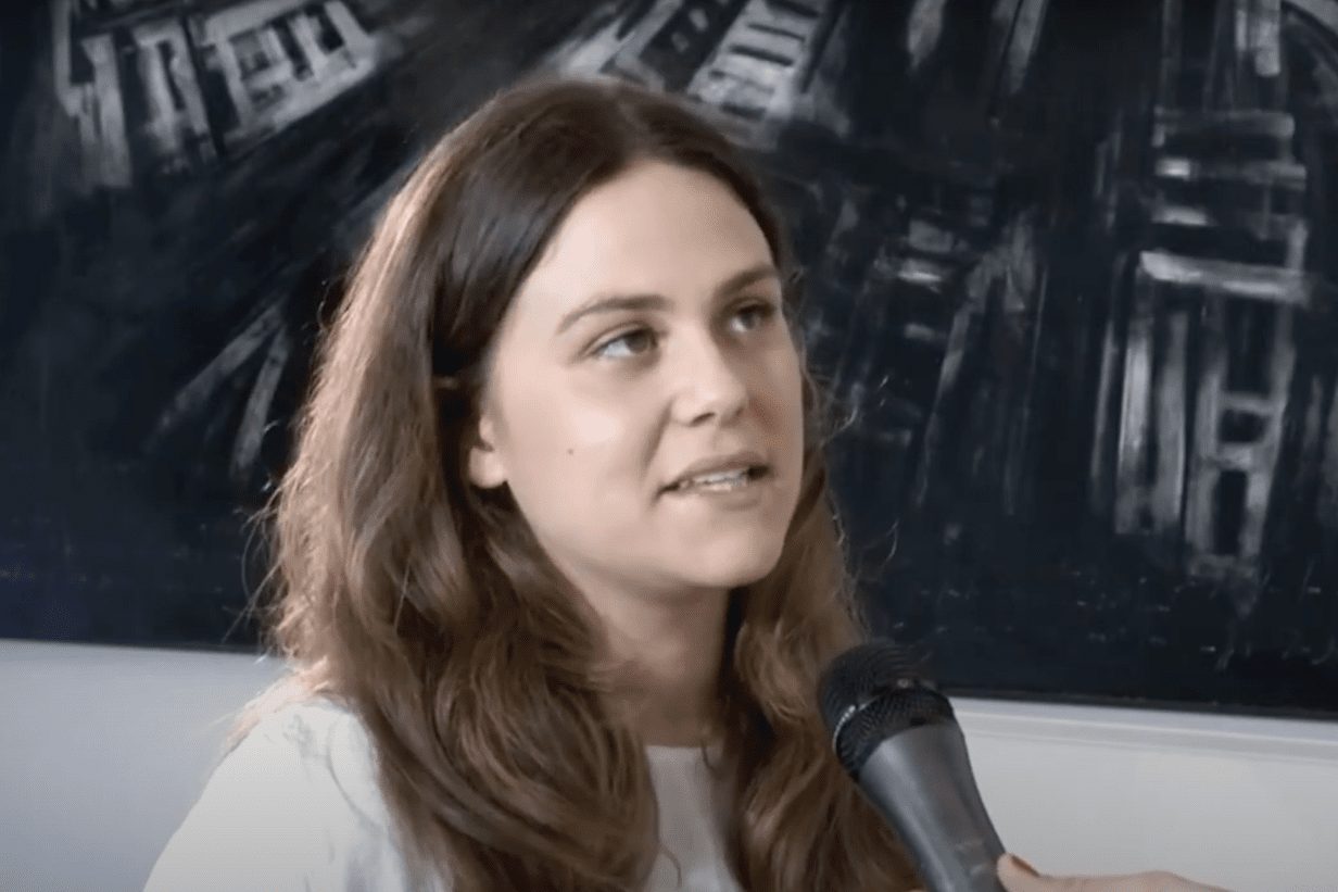 A woman speaking into a handheld microphone in front of a dark painted canvas, in a television interview frame